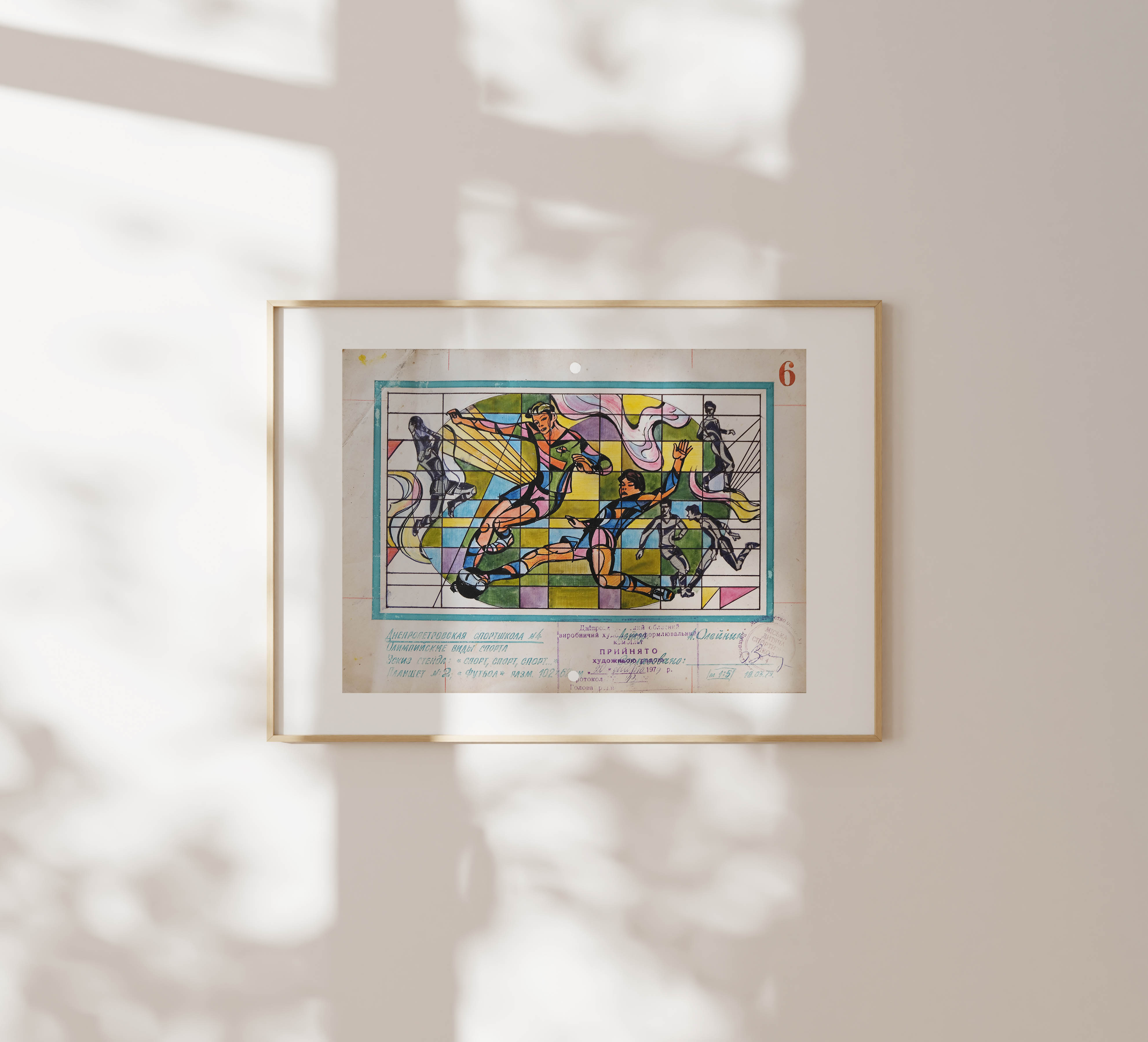 A framed drawing of soccer players hanging on a light-colored wall with shadows from leaves casting across.