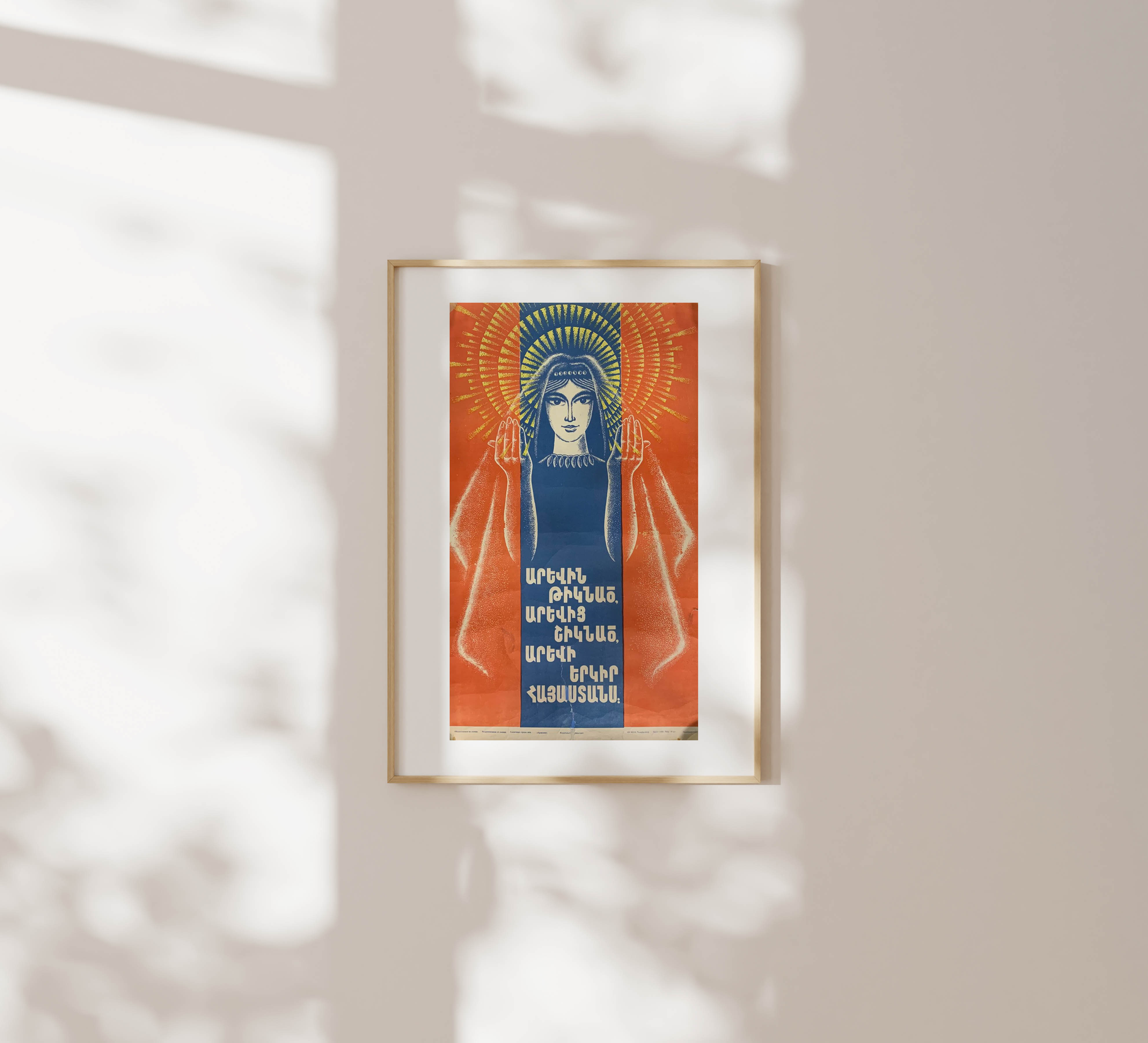Framed Soviet Armenian poster in a light wood frame hanging on a warm beige wall with leafy shadows cast across the surface.