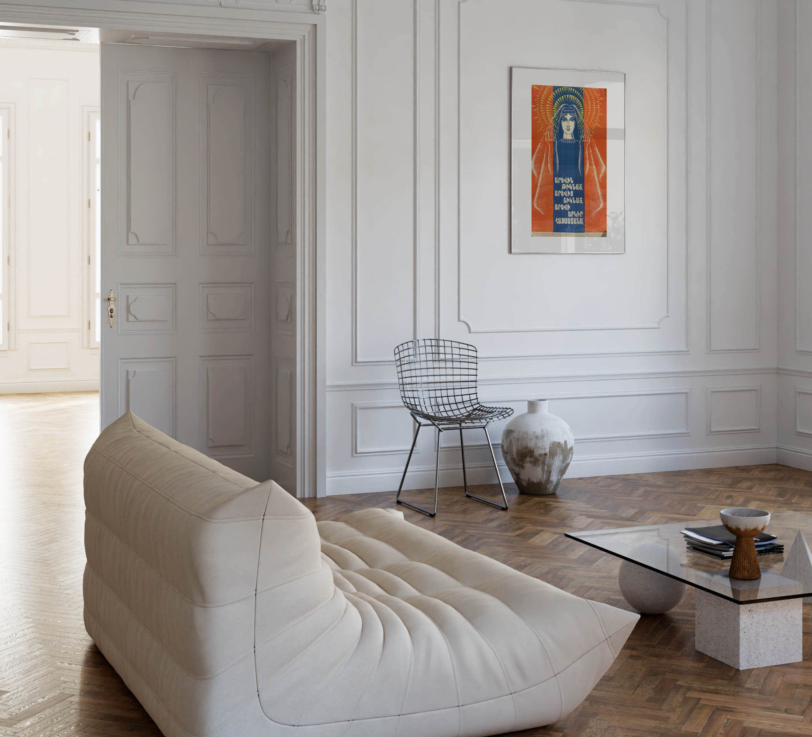 Bright white living room with a low cream-coloured modular sofa, a wire chair next to a rustic ceramic vase, and the framed Armenian poster hanging on a panelled wall.
